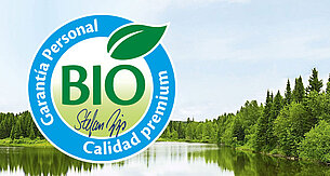 Un sereno paisaje de un lago rodeado de frondosos árboles verdes se superpone a un logotipo circular azul en el que se lee "BIO" en verde, "Stefan Hipp" en blanco y "Con garantía personal" en azul.