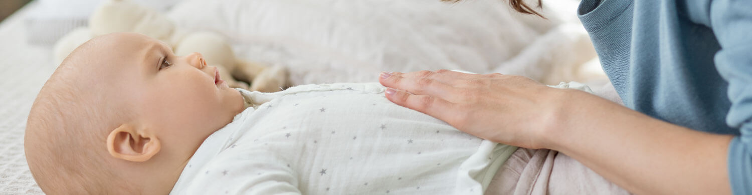 A close-up of a baby's face, looking up, with a hand gently placed on the baby's chest.