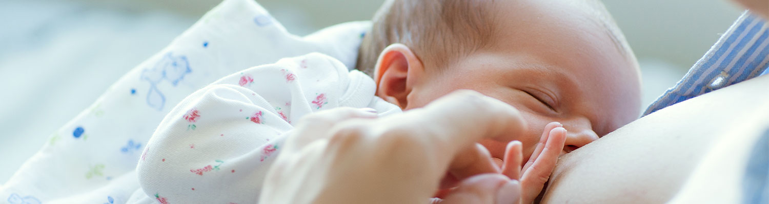 A close-up of a baby's face, partially obscured by a hand, with a white cloth featuring small floral patterns draped over the baby's shoulder.