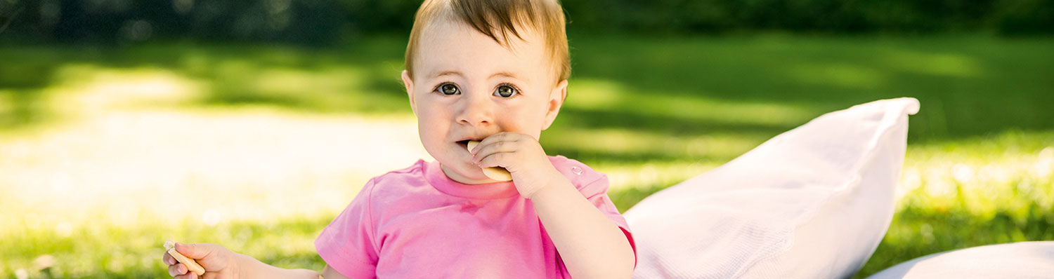 A baby in a pink shirt is seated outdoors on a blanket, holding a small object in one hand and a biscuit in the other, with a white pillow beside them.