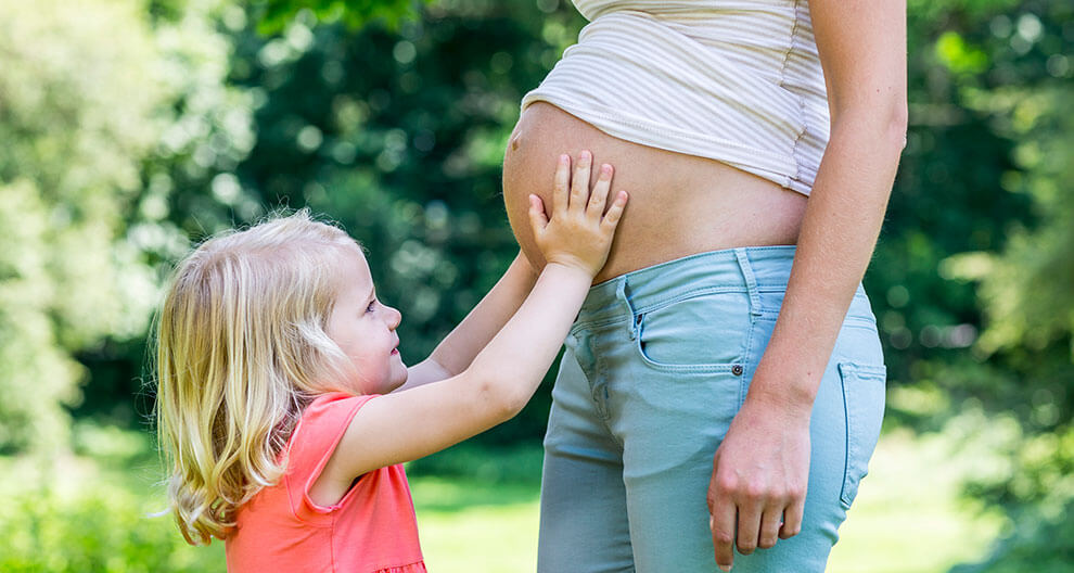 A young girl with blonde hair, wearing a pink shirt, places her hand on the belly of a taller person wearing a striped shirt and blue jeans.