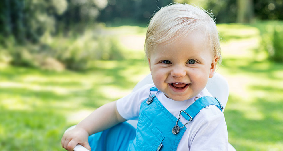 A toddler with blonde hair and blue eyes is sitting outdoors, wearing a white shirt and blue overalls, with a white object in their hand.