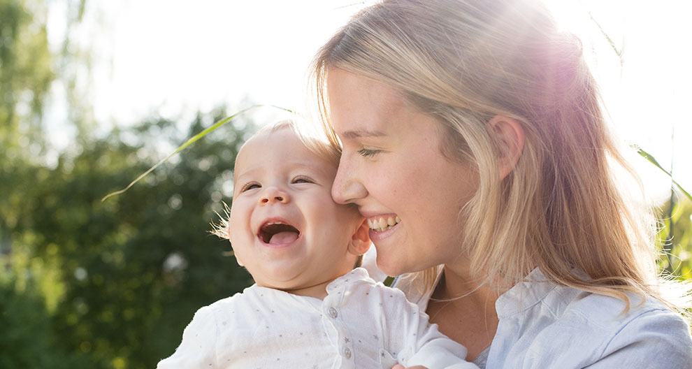 A woman and a baby are outdoors, with the woman holding the baby close to her face, and the baby is laughing.