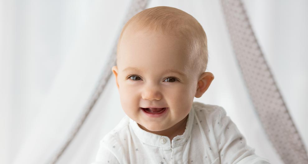 A baby with a white outfit and a dotted curtain in the background.