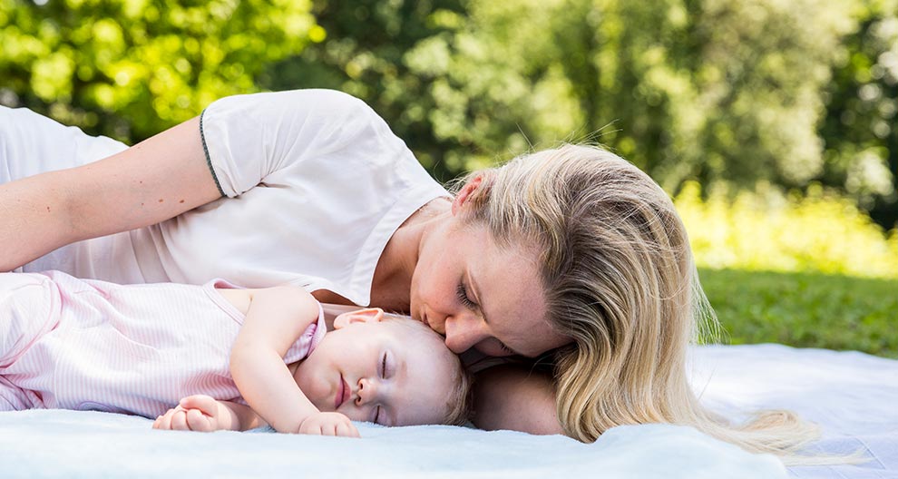 A woman in a white shirt is lying on a blanket outdoors, holding a sleeping baby dressed in pink.