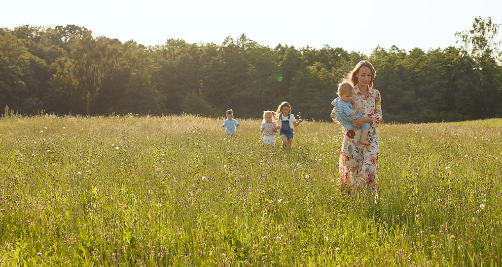 A woman in a floral dress holds a child while three other children run behind her in a grassy field with trees in the background.
