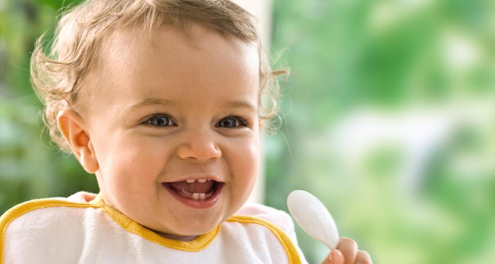 A baby with curly hair, wearing a white bib with yellow trim, holding a white spoon, smiling with a tooth missing.