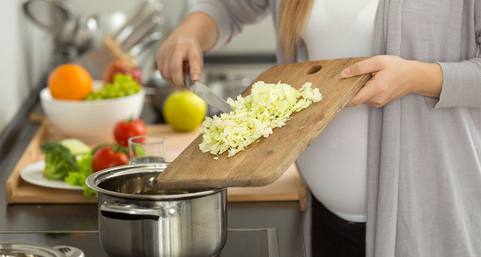 A person is chopping cabbage on a cutting board, with a pot on the stove and various vegetables and fruits in the background.
