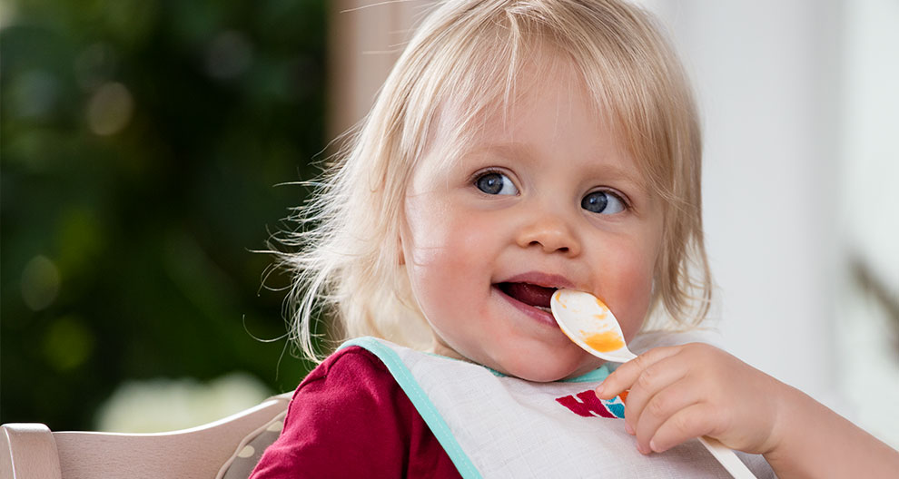 A young child with blonde hair is eating with a spoon.