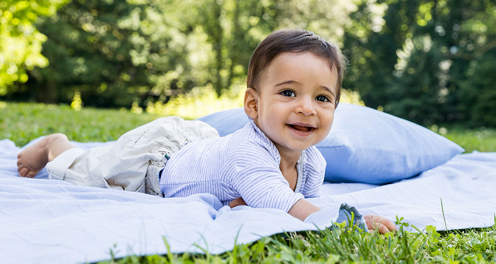A baby lies on a blanket in a grassy area with trees in the background.