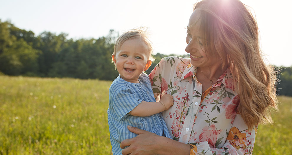 A woman in a floral blouse holds a laughing baby in a blue striped shirt, both outdoors with a green field and trees in the background.