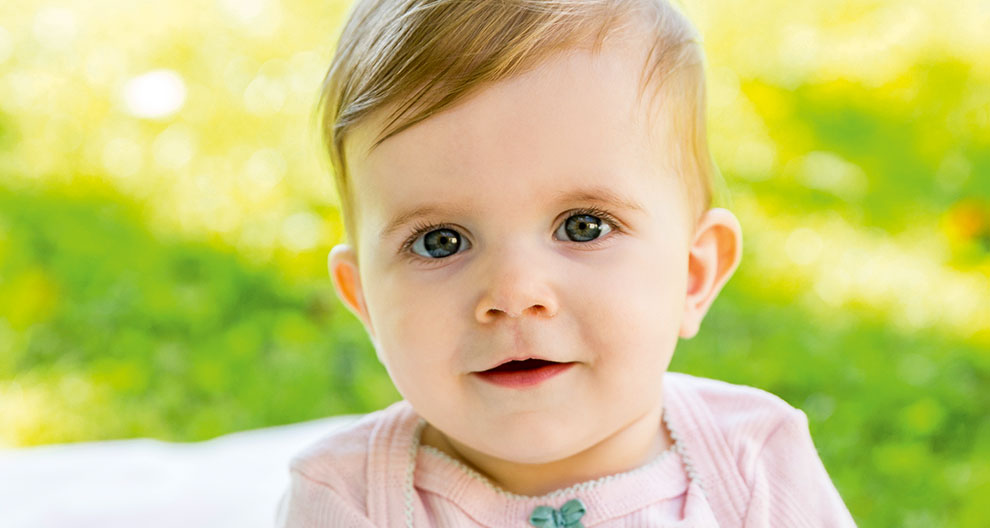 A baby with blue eyes and a pink outfit is sitting outdoors on a blanket.