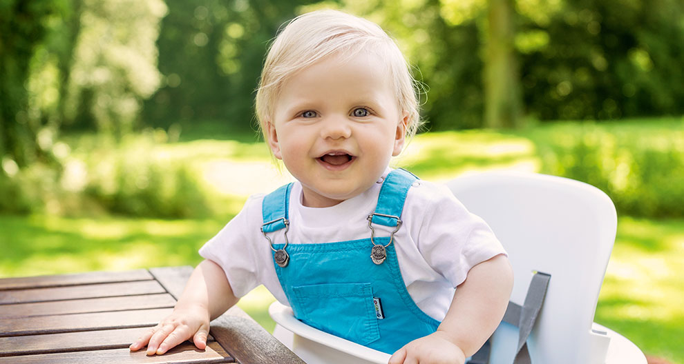 A toddler in blue overalls sits in a white high chair outdoors.