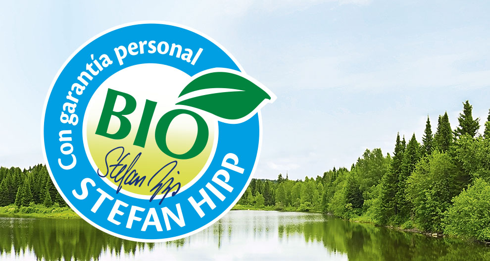 Un sereno paisaje de un lago rodeado de frondosos árboles verdes se superpone a un logotipo circular azul en el que se lee "BIO" en verde, "Stefan Hipp" en blanco y "Con garantía personal" en azul.