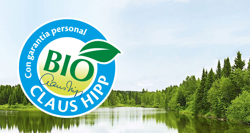 Un sereno paisaje de un lago rodeado de densos árboles verdes se superpone a un logotipo circular azul en el que se lee "BIO" en blanco, "Con garantía personal" en texto más pequeño y "CLAUS HIPP" en letras más grandes.