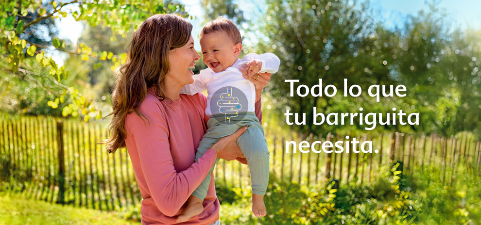 Una mujer con camisa rosa sostiene a un bebé risueño con camisa blanca y pantalones verdes, con una valla y árboles de fondo.